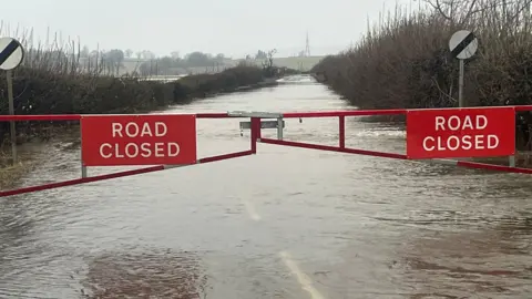 A flooded country road which has been sealed off behind a gate with two red signs reading 'road closed'