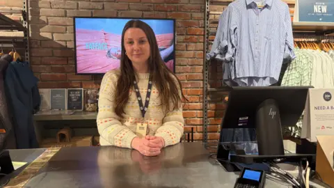 Nancy is stood behind the till in FatFace and wears a white jumper. Behind her is shirts on rails with a tv that reads FatFace. The desk which holds the till is silver.