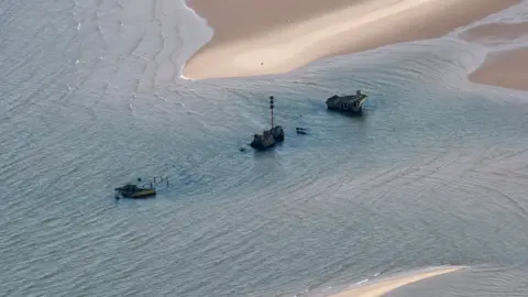 John Fielding Aerial Images Three sections of black shipwreck surrounded by sea water