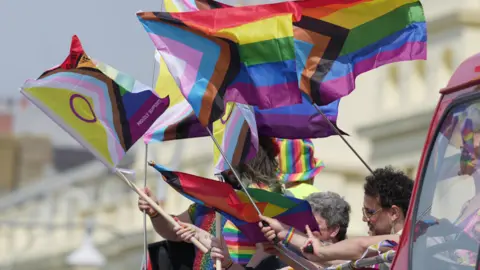 Eddie Mitchell People onboard an open top bus waving rainbow and trans pride flags