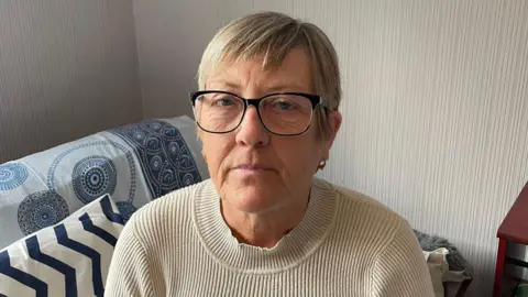 A woman in her 60s with short blonde hair. She is wearing black framed glasses and a beige coloured turtle neck jumper. She's sitting on her sofa with a patterned throw and cushion behind her. She's looking at the camera with a serious look on her face.