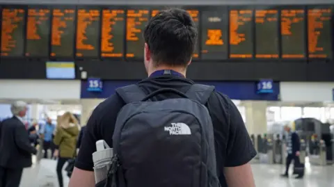 A man with a bag on his back is standing in front of electronic boards showing rail information at a station. 