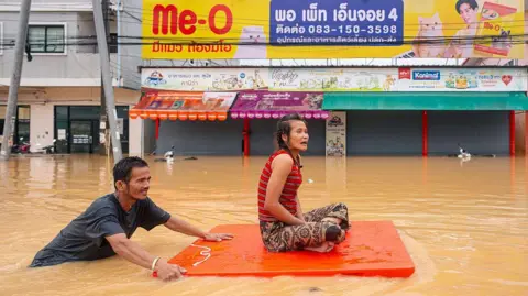 Getty Images Een man vervoert een vrouw op een oranje plastic bord door overstromingswater in Hat Yai in de zuidelijke provincie Songkhla in Thailand