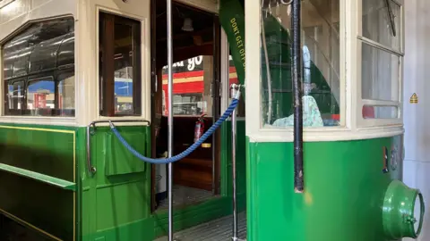 GUY CAMPBELL/BBC The entrance to a green and white tram with a silver metal rod down the centre of the gangway and a blue cord hanging up over the entrance