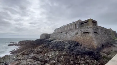 A large stone coastal castle sits on a rocky shoreline beside rough, grey-blue sea. Thick defensive walls with small rectangular openings run along the structure, and a long building with dormer windows stretches across the top. Waves gently lap against the rocks below under an overcast, cloudy sky.