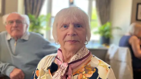 Jan is wearing a floral dress with a pink silk scarf wrapped around her neck. 