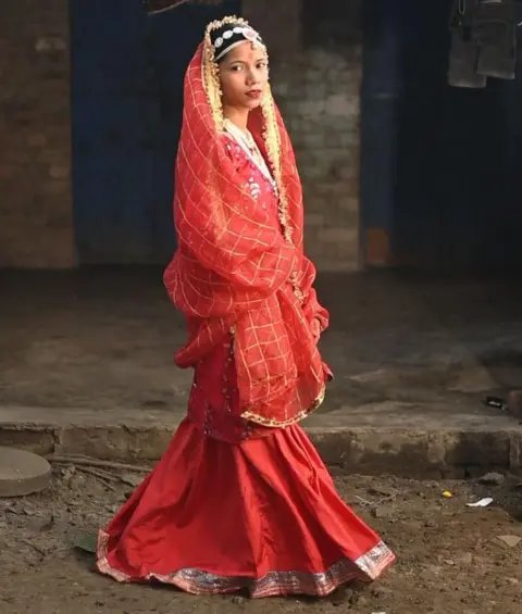 Innovation for Change A girl posing for the camera wearing colorful red clothes and jewelry stands outdoors on the street and looks directly at the camera. The bright lighting makes the costumes look vivid. 