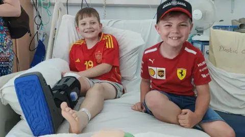 Jo Newman Theo Rees visited by his best friend Jacob, with both sitting on a hospital bed. Both are wearing red t-shirts and shorts and are smiling. Theo is wearing a big black and blue boot on one foot.