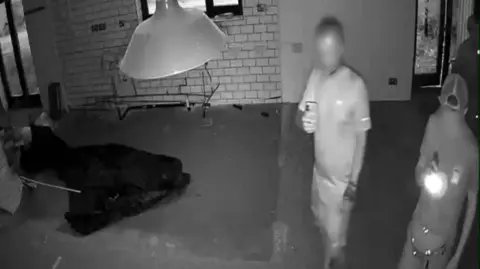 A black and white CCTV image of two men in an abandoned building. One is taking photos on his phone and the other appears to be shining a torch. 