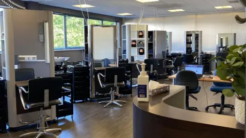 Poultec Training View of a hairdressing salon. A row of four chairs is facing large mirrors. Each base has a trolley full of hairdressing supplies on it. There are some model heads with synthetic hair for the students to practise on. There is a reception desk and a computer. 