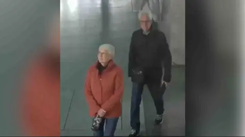 British Transport Police Woman in orange jacket and man in black jacket, both wearing jeans and both with grey hair. They are walking across a paved or tiled floor.