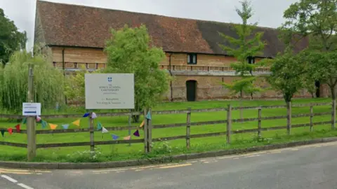 A Google streetview image of the Junior King's School in Canterbury. The image shows a long building with a grassy field in front. There is a wooden fence and a sign that reads "Kings School Canterbury"