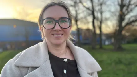 Katarzyna Wysokinska, who has large black framed glasses and blonde hair tied back, and is wearing a light grey jacket over a black top, smiles into the camera against a backdrop of a grassy area dotted with trees. 