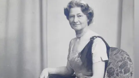 A black and white photo of a lady sat in an arm chair with a long dress on