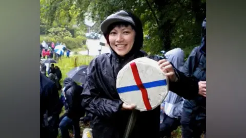 Billy Kidd A woman wearing a black plastic raincoat holding a wheel of cheese and smiling