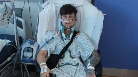 Liam McCracken Liam McCracken, who is wearing a patterned hospital gown, is sitting in a chair next to a hospital bed. He is wearing a neck brace and is surrounded by medical equipment. 