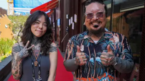 Two people with tattoos smile for the camera while standing on a red carpet