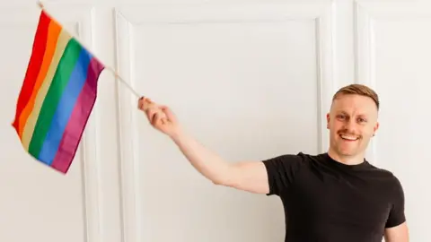 Ian Timbrell Ian Timbrell with short, fair hair wearing a black short-sleeved T-shirt holds up a small rainbow flag in his right hand, standing in front of a white, panelled wall.