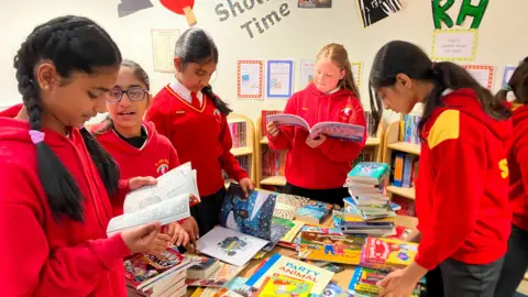 A group of girls in a primary school stand around a table piled high with books. They all wear red hoodies.