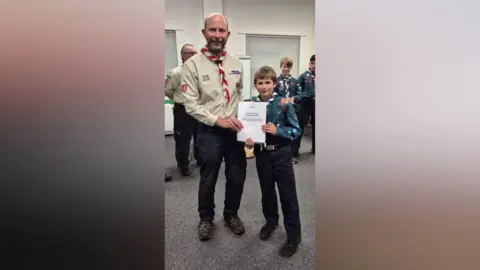 Jersey Scouts Glen and Charlie smile at the camera as they hold a certificate between them. They're both wearing Scouts uniforms and there a few Scouts behind them. Glen has a cream coloured shirt with a red and white neckerchief. Glen has a stubbly beard and a bald head.