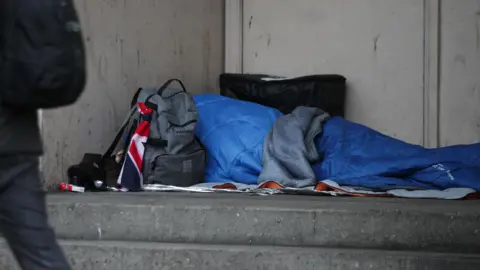 A homeless person sleeping rough in a doorway. They are in a blue sleeping bag with their belongings around them in other bags