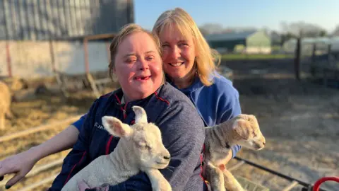 Lucy Best and her mum Jo Best sitting side by side. Lucy is on the left and is wearing a grey jumper. Her mum is wearing a light blue jumper. They are both smiling and holding lambs. There is a farm yard in the background. 