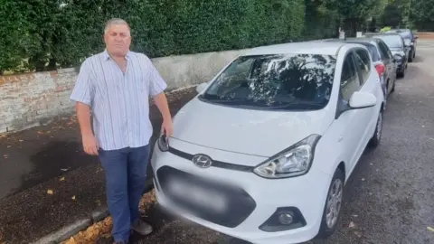 Richard Lynn A man stands by a white parked car