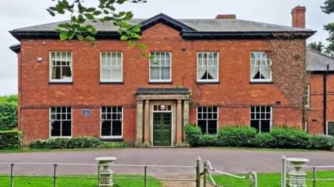 Ian Hughes A large red-brick building with columns on either side of the front door and large paned windows, with a drive and a fence in front of it