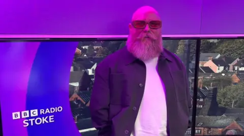 A man in glasses, with a bushy beard and curly moustache is smiling at the camera. Behind him is a graphic showing houses, and BBC Radio Stoke branding