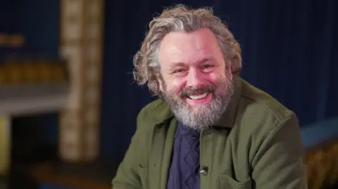 Michael Sheen sits in a theatre looking at the camera. He has long wavy greying hair and a beard. He is wearing a green jacket and blue jumper. 