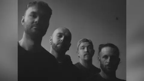 The four band members of Collapse The Sky face the camera, all of them unsmiling. They are photographed in a black and white and one of the them is wearing a cap. 