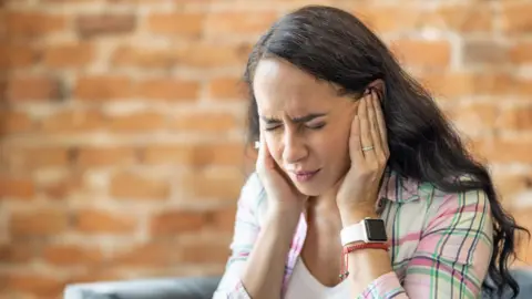 A woman with dark hair covering her ears, she is wearing a check white pink green and navy shirt