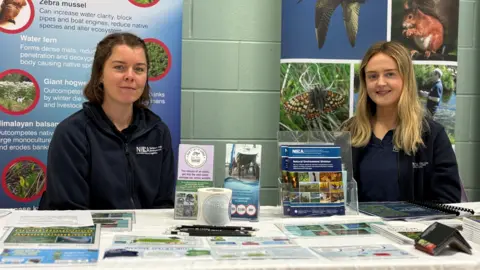 Two women sit at an information stall desk with leaflets on the table in front of them. They wear blue hoodies. The woman on the left has brown hair and the woman on the right has blonde hair. There are posters behind them with photos of wildlife.