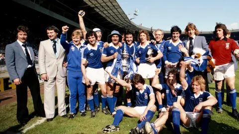 Allsport/Getty Images The FA Cup winning Ipswich Town team from 1977/78. Many of the players are shouting in joy as they smile at the camera. Some have their arms in the air. 