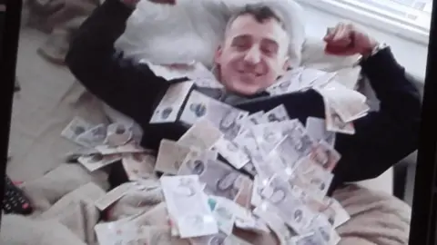 Humberside Police Thomas Crombie lying on a bed and covered in cash. He has his arms up and is smiling at the camera. He has short brown hair and is wearing a black jumper.