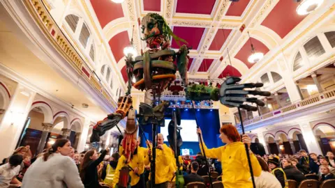 Tom Arran Several people wearing yellow fleeces man a large puppet inside Hull City Hall, children surround the puppet and the photo is taken from below to show the size of the creature.