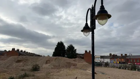 Martin Heath/BBC A landscape of light-coloured soil with a mound in the centre. A few green bushes have sprouted up on the ground. There is a lamp standard with two dewdrop lamps at the top. A street of shops can be seen on the other side of the road to the right.