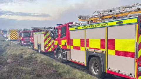 A row of fire appliances parked by the side of a moorland road. The vehicles are brightly painted yellow and red. Grey smoke drifts overhead.