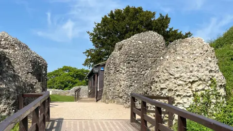 View from a footbridge going into ruins and a visitors wooden hut. Green grass, trees and blue skies.