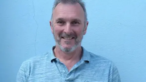 MANX DEAF SOCIETY Nigel Latham, a man with grey hair and a stubble who has a friendly smile, he wears a light blue T-Shirt and stands in front of a blue wall.