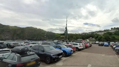 Google Cars parked in a row on the pier. behind them are railings and the quay. Grass covered cliffs are behind the water. The statue is in the centre. The statue depicts a pregnant woman holding aloft a sword while carrying the scales of justice and standing on a pile of law books