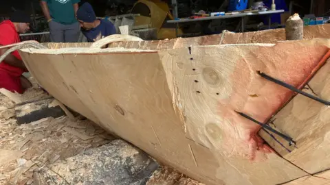 A partially carved log begins to take the shape of a canoe. Three carvers are pictured at the far end of the log
