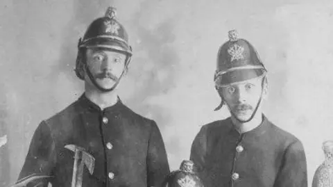 Family Handout An old black and white photograph, showing two men facing the camera.
On the left a taller man wearing a buttoned coat and old fire service helmet. He has a large moustache and can be seen holding an axe in his right hand. On the right, also in uniform is a man wearing similar attire, looking at the camera with a straight face. 