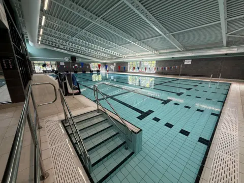 A swimming pool, with steps in the foreground and flags at either end. There is a white metal ceiling with several beams stretching the width of the pool room. There is railing on either side of the steps and windows in the distance.