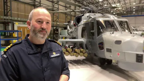Commander Andrew Henderson stands next to a wildcat helicopter in a large hangar. He is wearing a navy uniform.