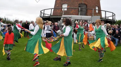 Trustees of Thaxted Windmill Female Morris dancers wearing white and green dance around the base of the windmill as a crowd watches on