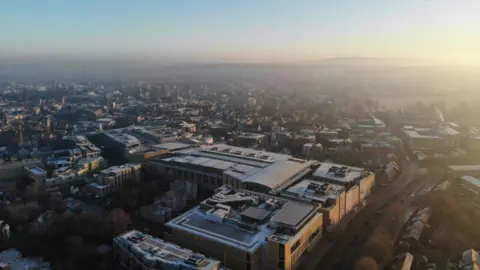 An aerial view of Oxford city on a winter morning. Spires are in the far distance, but it is mainly modern-looking buildings in the foreground.