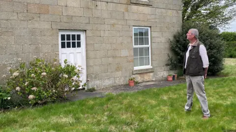 BBC Martin Beal is looking at his house which is a Georgian stone built farmhouse with a white door and windows. He is a man, about 60 years of age, wearing a waist coat, white shirt and light coloured trousers.