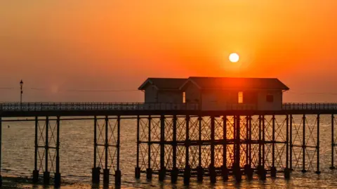 Nadezna / Weather Watchers Wooden pier and buildings on the end in orange evening sun which glows bright above 