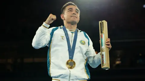 Reuters Hasanboy Dusmatov raises his fist in the air and celebrates the Olympic medal for the Paris 2024 Games around his neck. He wears a white tracksuit with blue and gold stripes